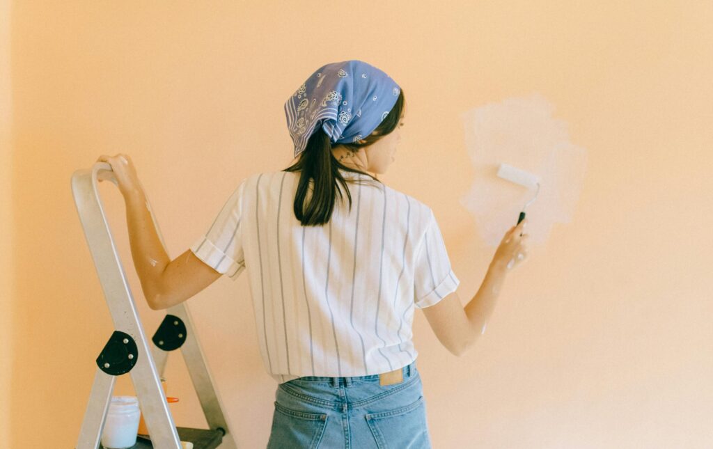 A woman with a bandana using a paint roller to paint a peach-colored wall indoors.